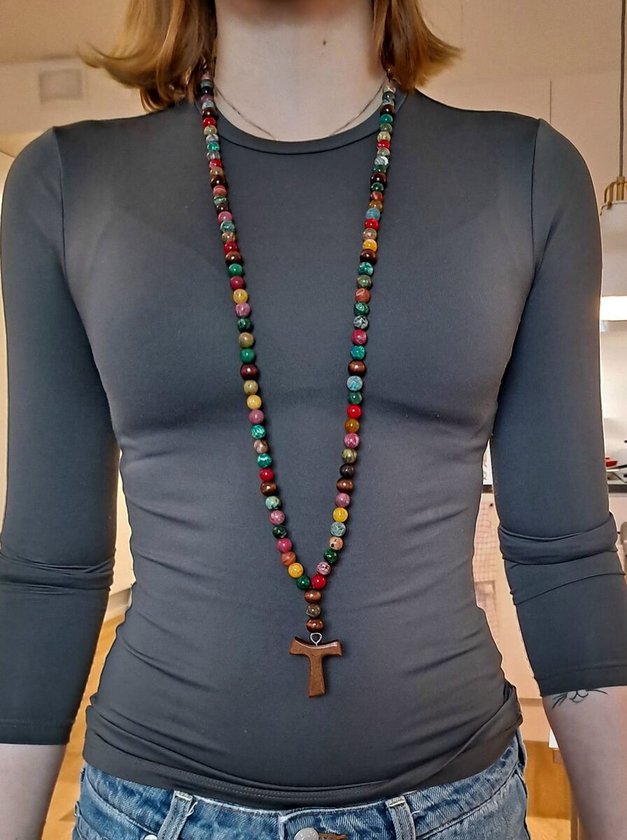 Pitkä, värikkäistä kivihelmistä tehty protestanttinen rukousnauha kaulakoru, jossa puinen TAU-risti, mallin kaulassa harmaan paidan päällä.