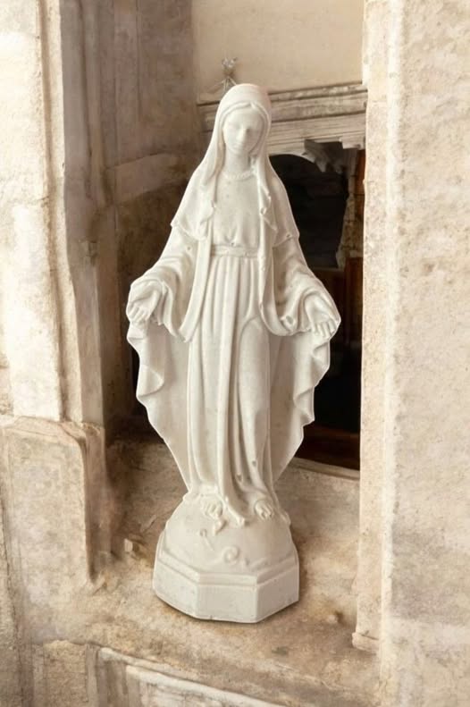 Valkoinen Äiti Maria -patsas, joka seisoo kivisessä ikkunasyvennyksessä, kädet ojennettuina siunauksen asennossa.