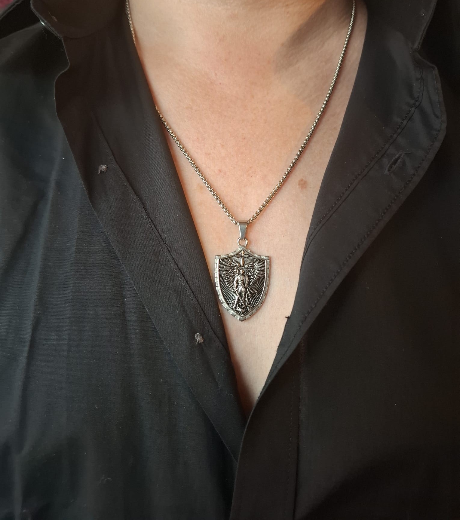 Lähikuva miehen kaulasta ja ylävartalosta. Hänellä on musta paita, ja kaulassa roikkuu hopeansävyinen teräskoru, jossa on kilven muotoinen riipus ja keskellä arkkienkeli Mikael.