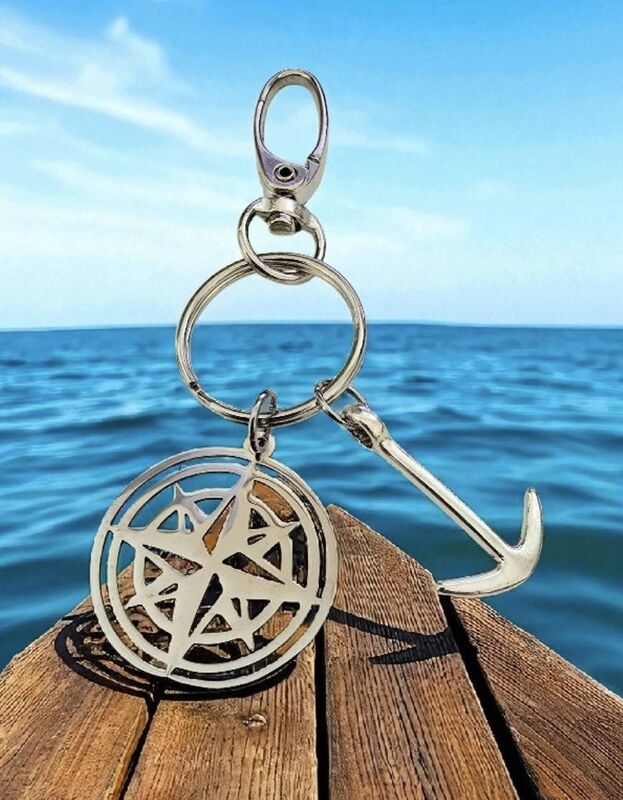 Hopeinen ankkuri- ja kompassiriipuksella varustettu avaimenperä puulaiturilla meren rannalla kirkkaan sinistä taivasta vasten.