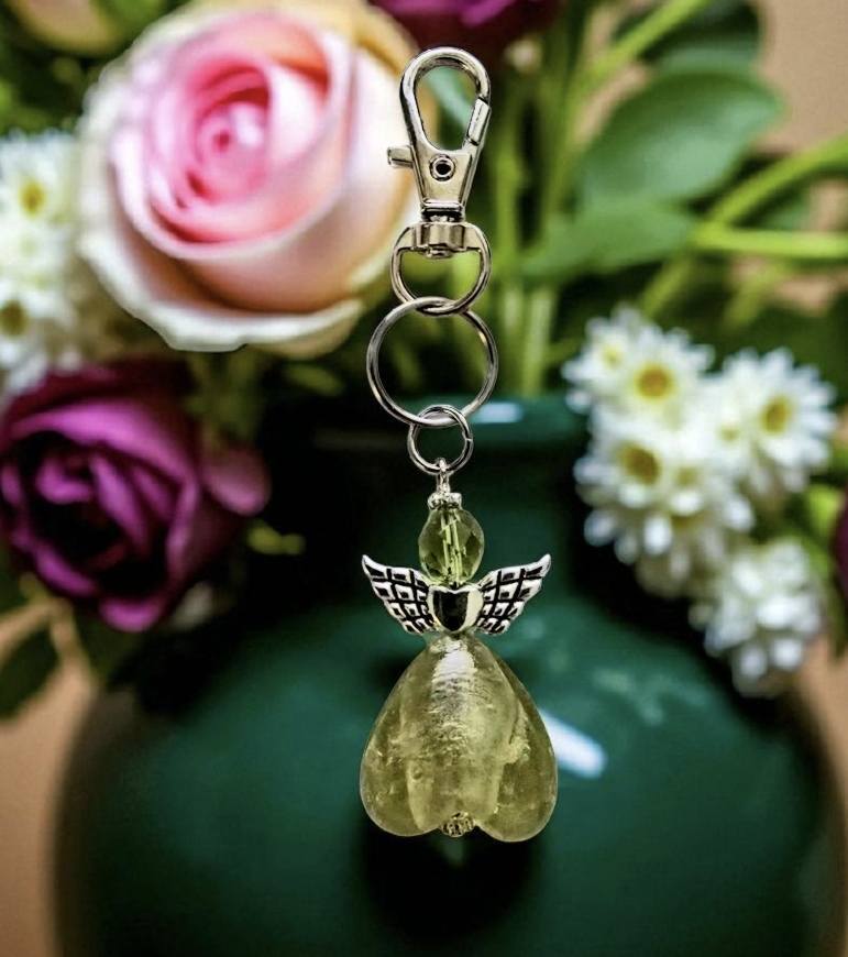 Vaalean oliivinvihreä enkelilaukkukoru, jossa metalliset siivet ja suuri karabiinilukko, taustalla värikkäitä kukkia.