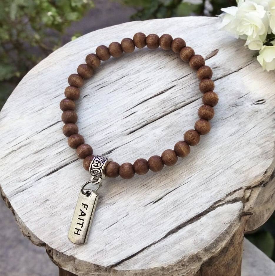 Puinen helmirannekoru metalliriipuksella, jossa lukee FAITH. Koru on aseteltu vaalealle puulle.