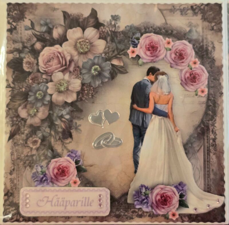 Hääkortti, jossa morsiuspari seisoo yhdessä sydänkehyksen edessä. Kortissa on vaaleanpunaisia ja violetteja kukkia sekä hopeiset sydän- ja vihkisormuskoristeet. Teksti: 'Hääparille'.