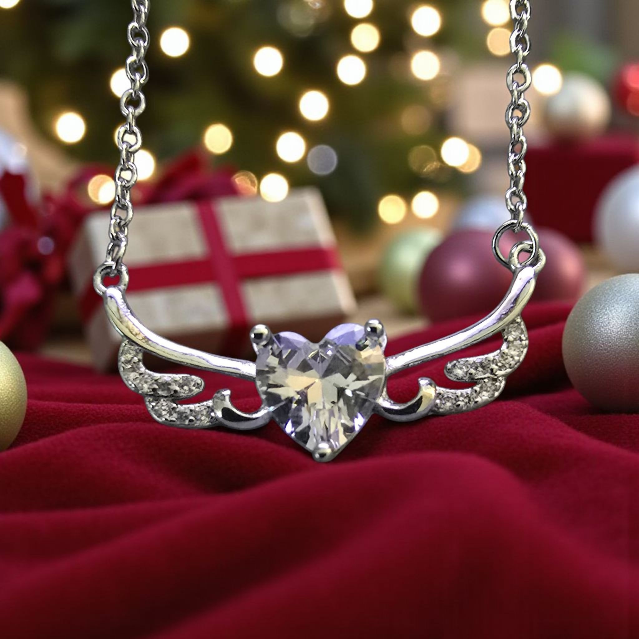 Hopeinen kaulakoru, jossa kirkas sydänriipus ja siipimäinen koriste. Taustalla näkyy joulukuusen valoja, lahjapaketteja ja koristepalloja.