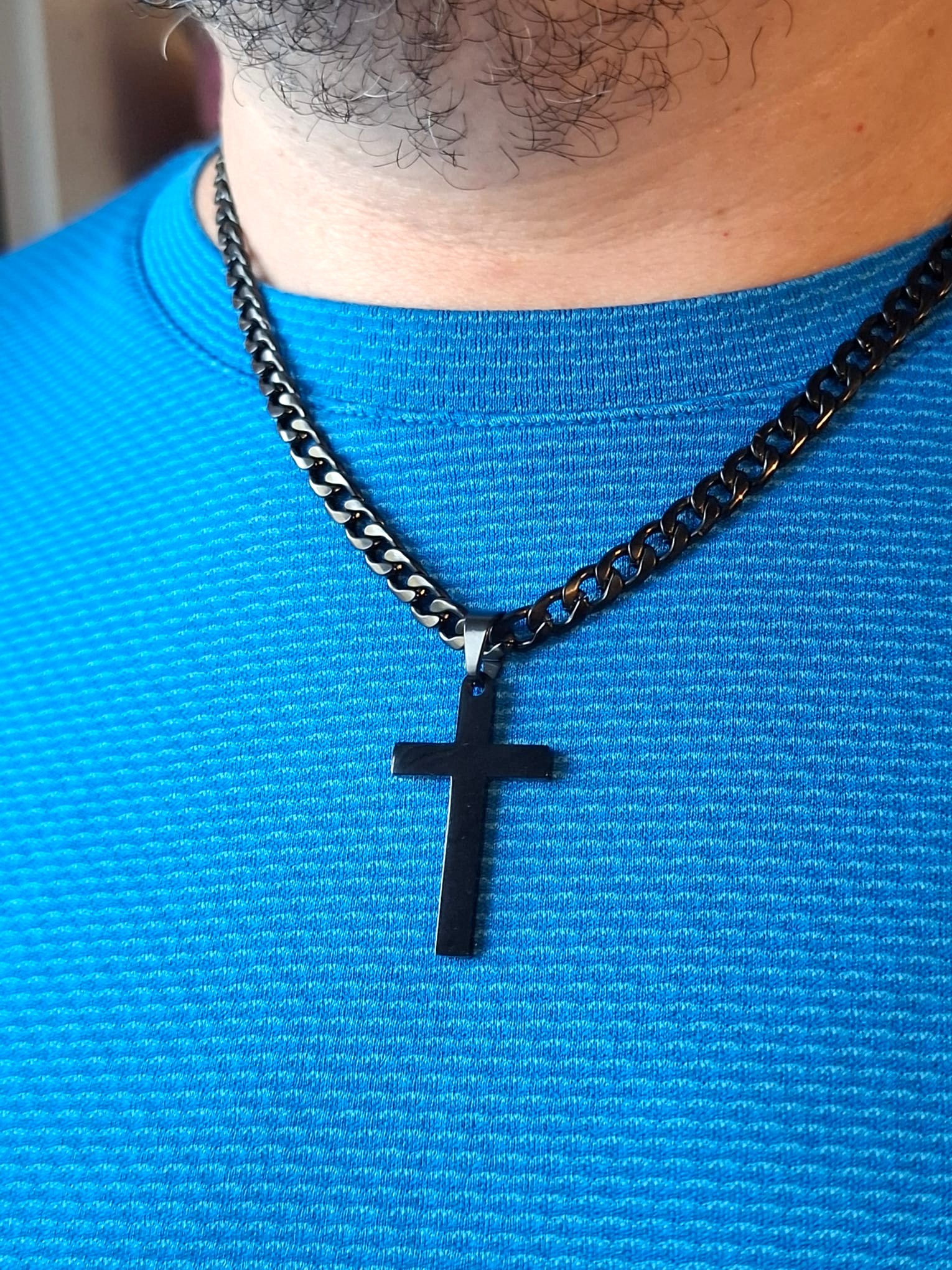 Lähikuva miehen kaulasta ja ylävartalosta. Hänellä on päällään kirkkaansininen paita ja kaulassaan leveä musta panssariketju, jossa roikkuu musta risti. Kuvassa näkyy myös hieman partaa ja kaulan ihon yksityiskohtia.