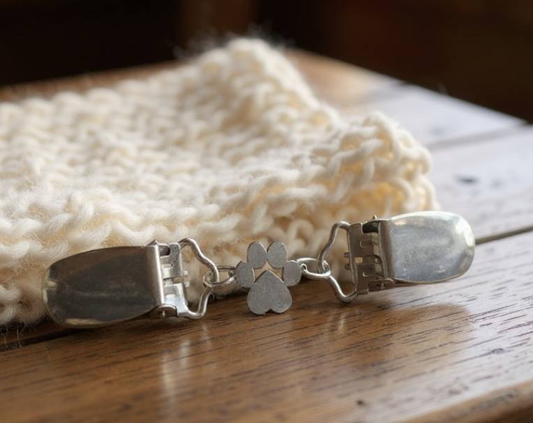 Hopeinen neuleklipsi tassuaiheisella koristeella, aseteltuna valkoisen villahuivin viereen puupöydällä.