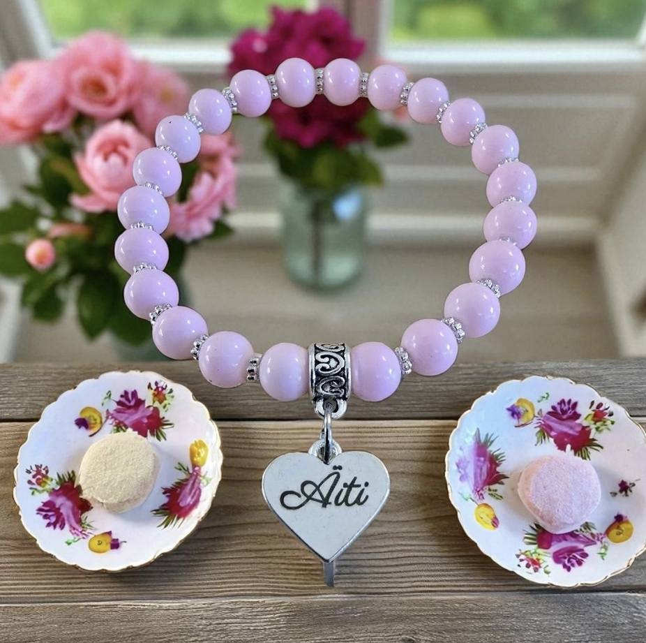 Vaaleanpunaisista helmistä käsityönä tehty rannekoru, jossa sydämenmuotoinen riipus tekstillä 'Äiti', taustalla kukkia ja kahvikupit.