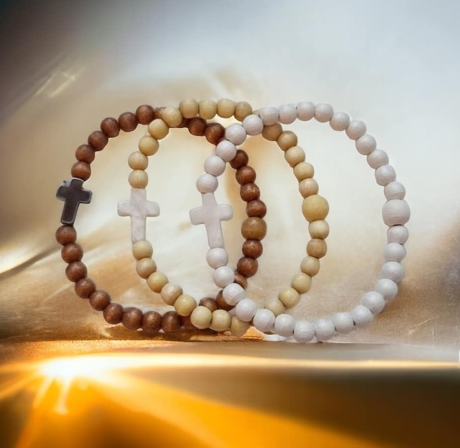 Kolme puuhelmistä käsityönä valmistettua rukousnauha rannekorua, joissa on kiviristi. Värit: ruskea, vaalea puu ja valkoinen. Taustalla lämmin, kullanhohtoinen valo.