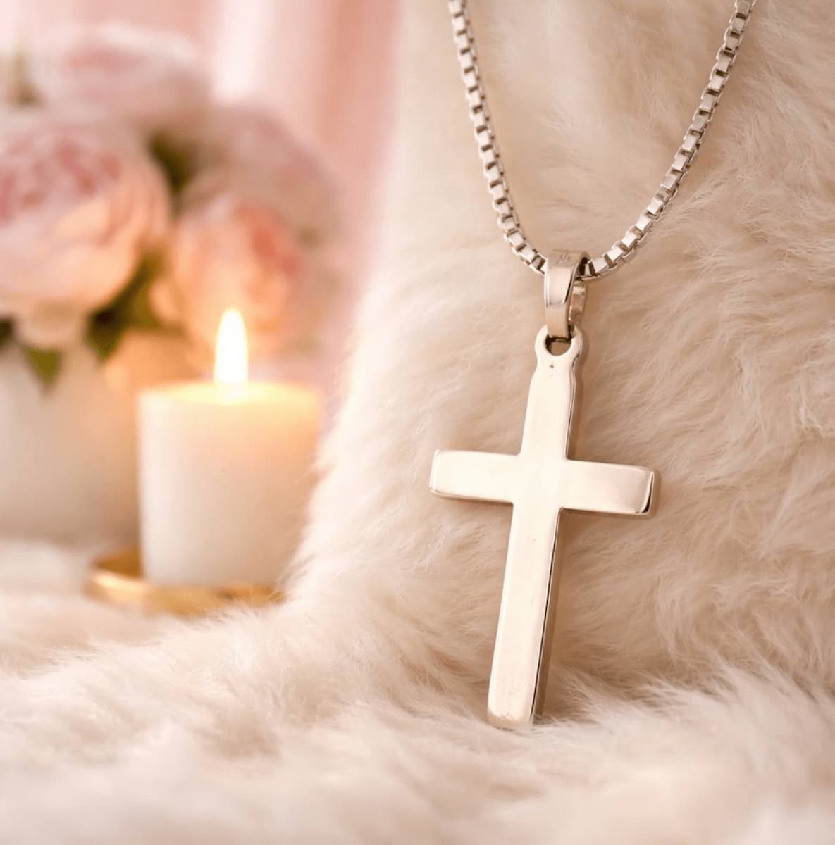 Hopeinen rippiristi kaulakorussa pehmeällä vaalealla taustalla, risti roikkuu hopeaketjussa ja taustalla näkyy kynttilä ja vaaleita kukkia.