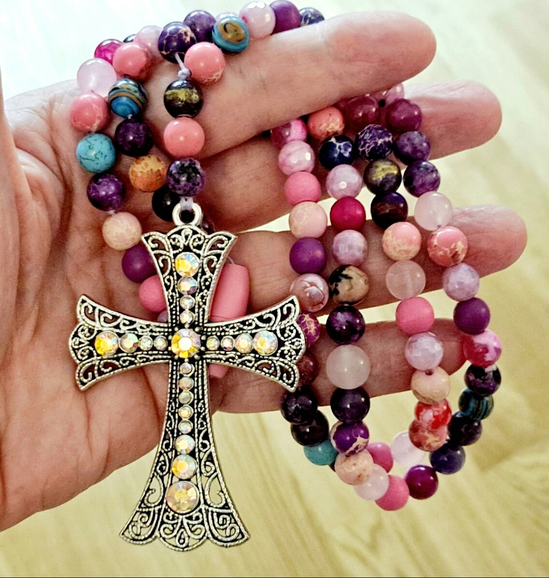 Käsi pitelee värikästä kivihelmistä tehtyä rukousnauhaa, jossa on suuri strassikoristeinen risti-riipus.