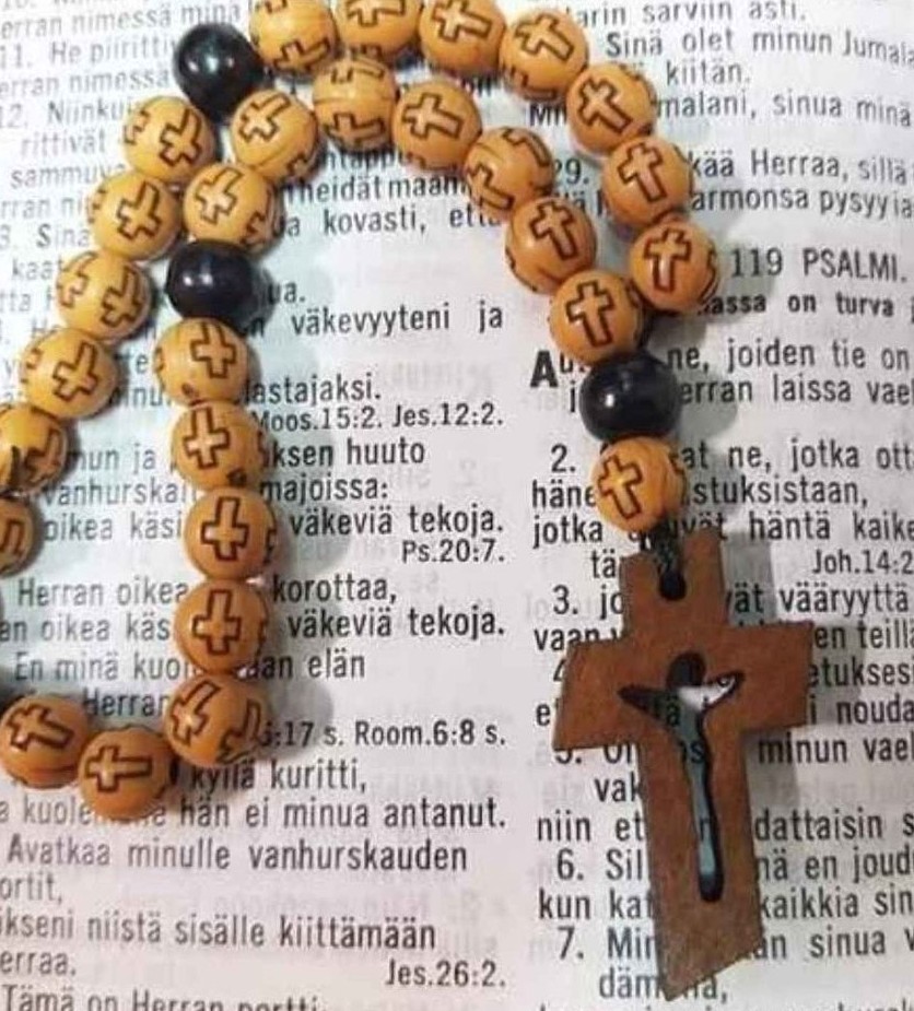 Akryylihelmistä tehdyt rukoushelmet, ristihelmikuvio ja puinen krusifiksi avoimen Raamatun sivun päällä.